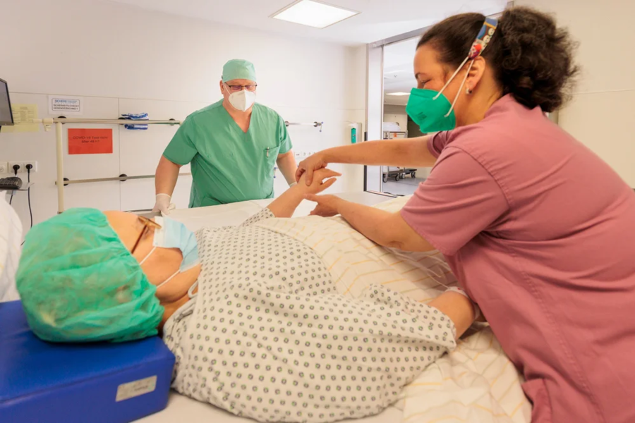 Krankenpflegerin misst Puls eines Patienten auf Krankenhausbett, Arzt im grünen OP-Kittel im Hintergrund