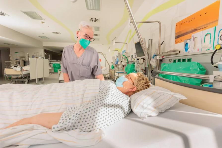 Krankenpflegerin steht neben einem Patientenbett in einem Krankenhauszimmer, Patient liegt mit Maske und Krankenhaushemd im Bett