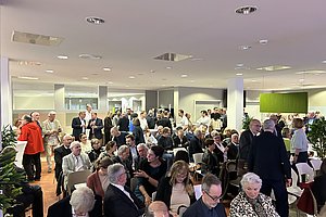 Große Gruppe älterer Menschen sitzt und steht in einem hellen Raum mit weißen Wänden und Deckenleuchten