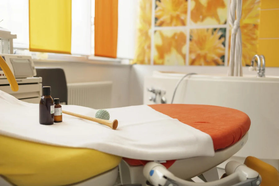 Ein Behandlungsstuhl mit gelber und orangefarbener Polsterung, auf dem eine weiße Decke liegt. Daneben stehen mehrere kleine Flaschen und ein Massagegerät.