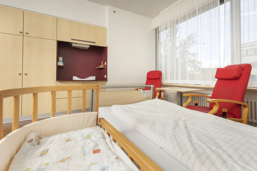 Ein helles Zimmer mit einem Holz-Kinderbett, einer weißen Bettdecke mit bunten Mustern und zwei roten Stühlen. Im Hintergrund sind Holzschränke und ein Fenster mit Vorhängen.