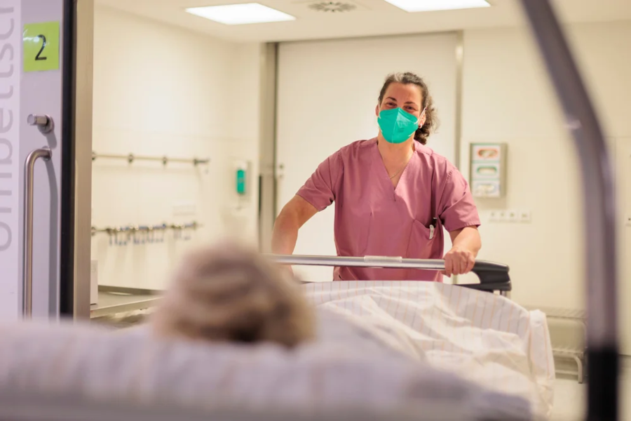 Pflegekraft in rosa Kittel schiebt Krankenhausbett mit Patient in einem hellen Krankenzimmer
