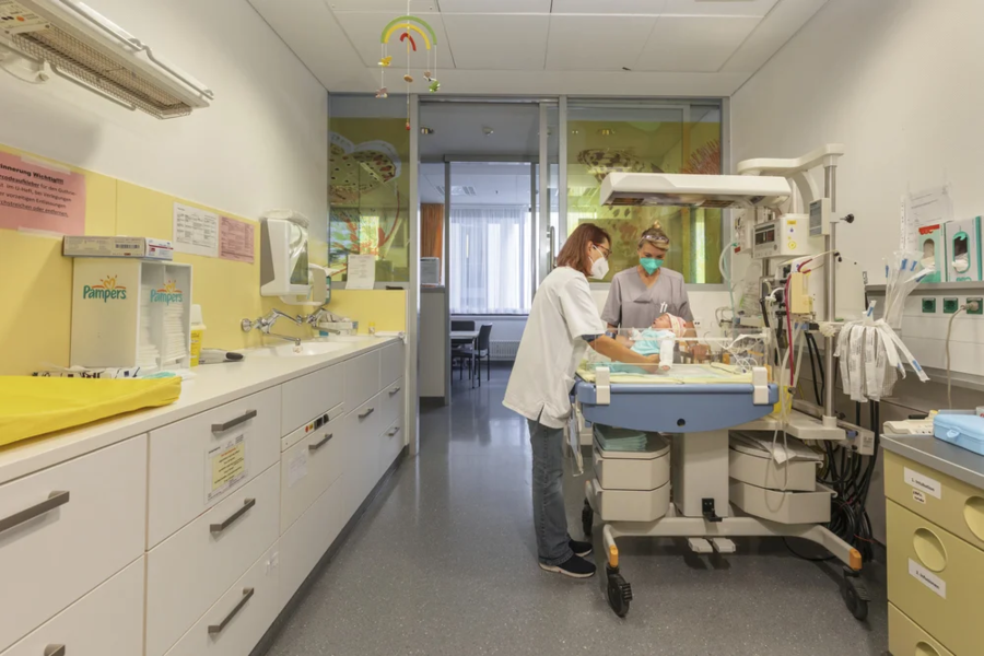 Zwei medizinische Fachkräfte betreuen ein Neugeborenes an einem Inkubator in einem Krankenhauszimmer