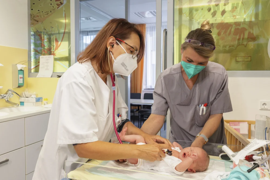 Zwei medizinische Fachkräfte in weißen Kitteln untersuchen ein liegendes Baby auf einer Liege. Eine Fachkraft hat ein Stethoskop um den Hals.