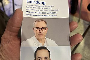 Hand hält Einladungskarte mit Text und zwei verpixelten Porträtfotos, im Hintergrund Stuhl und Tisch erkennbar