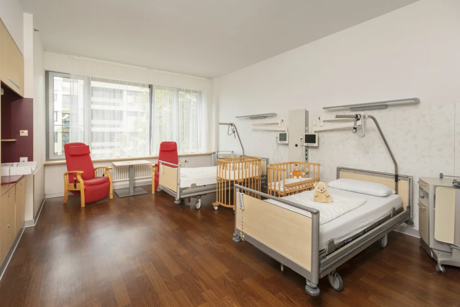 Ein Krankenhauszimmer mit zwei Betten, einem Kinderbett und einem roten Sessel. Auf dem Bett liegt ein Plüschtier. Es gibt medizinische Geräte an den Wänden.