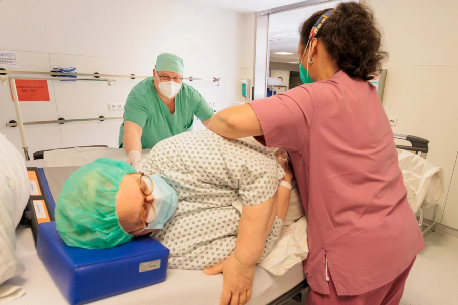 Pflegekraft in rosa Kleidung unterstützt Patientin in Krankenhauskleidung und grünem Haarnetz beim Umlagern auf einem Krankenhausbett