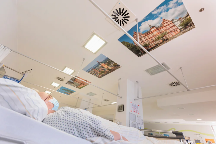 Ein Patient liegt in einem Krankenhausbett. An der Decke hängen Bilder von Gebäuden.