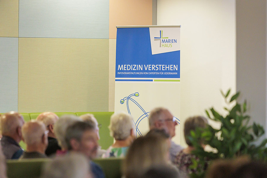 Publikum sitzt in einem Raum vor einem blauen Banner mit dem Text 'MARIEN HAUS MEDIZIN VERSTEHEN INFOVERANSTALTUNGEN VON EXPERTEN FÜR JEDERMANN'.