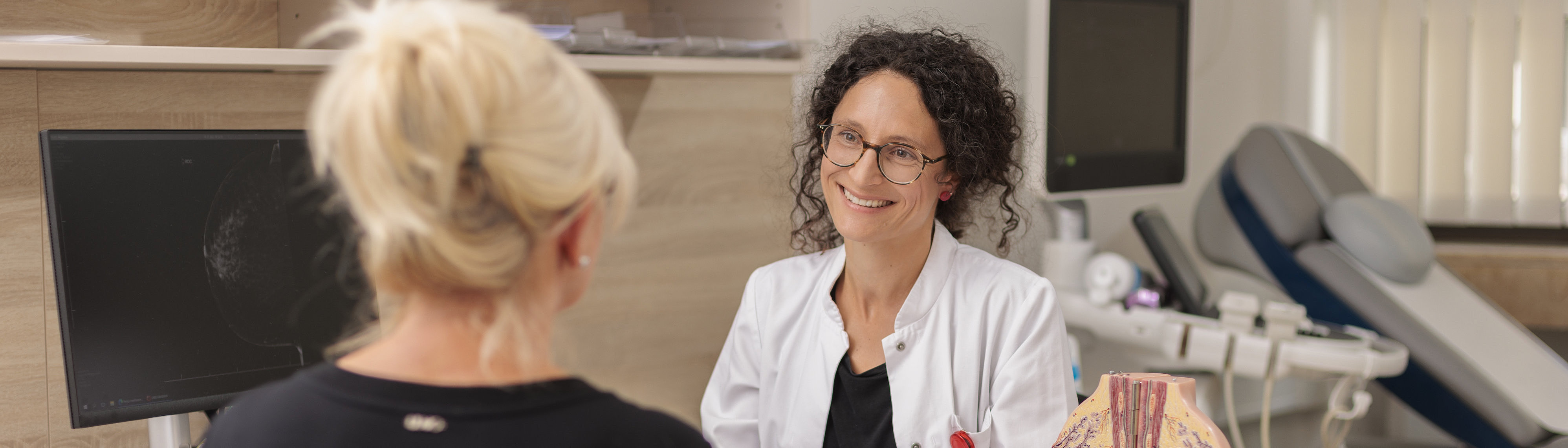 Ärztin im Gespräch mit Patientin in modernem Behandlungsraum mit medizinischer Modell-Darstellung.