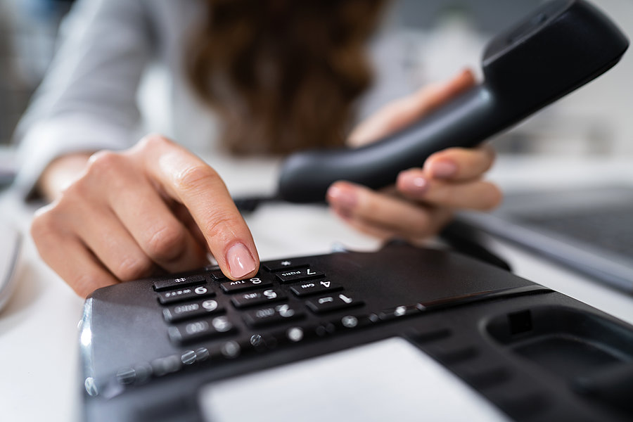 Eine Hand drückt eine Taste auf einem schwarzen Telefon, während die andere Hand den Hörer hält. Der Hintergrund ist unscharf.