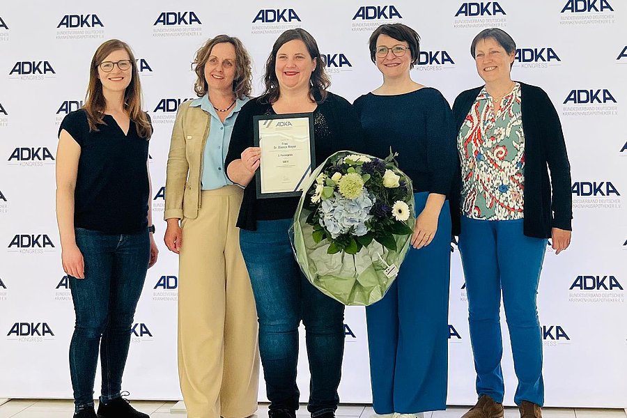 Fünf Frauen stehen vor einer Wand mit dem ADKA-Logo, eine hält eine Urkunde, eine andere einen großen Blumenstrauß.