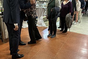 Menschen stehen in einer Schlange in einem Innenraum, vorne umarmen sich zwei Frauen, andere Personen warten geduldig.