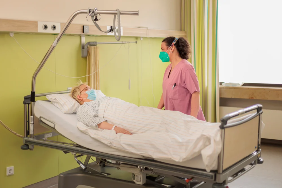 Krankenpflegerin in rosa Kittel steht neben einem Patientenbett in einem Krankenhauszimmer