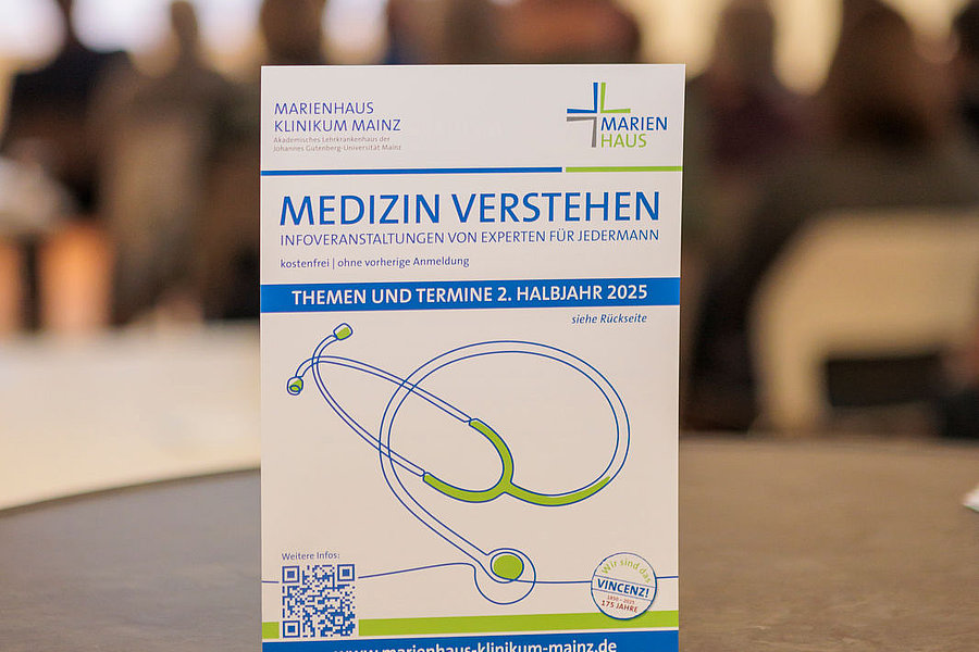 Ein Informationsflyer mit dem Titel 'MEDIZIN VERSTEHEN' und einem grafischen Stethoskop. Im Hintergrund sind unscharfe Personen zu sehen.