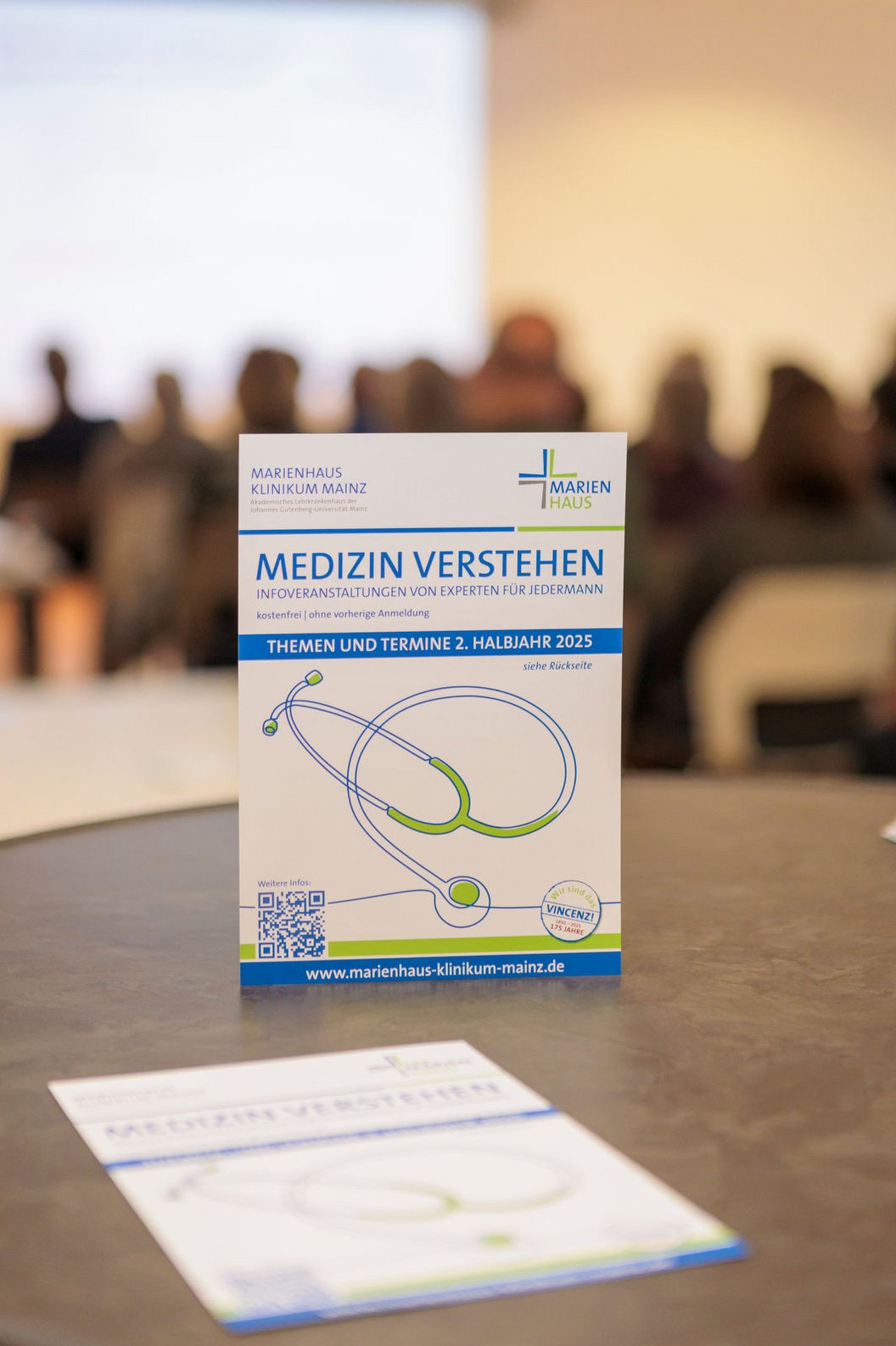 Ein Informationsflyer mit dem Titel 'MEDIZIN VERSTEHEN' und einem grafischen Stethoskop. Im Hintergrund sind unscharfe Personen zu sehen.