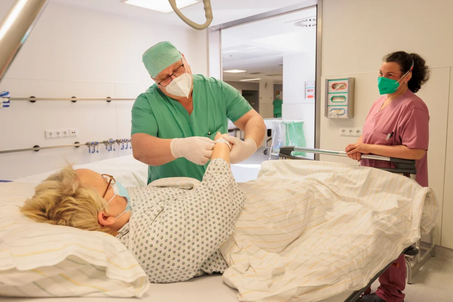 Krankenhauszimmer mit Patientin im Bett, Arzt in grüner OP-Kleidung untersucht ihren Arm, Pflegekraft steht daneben