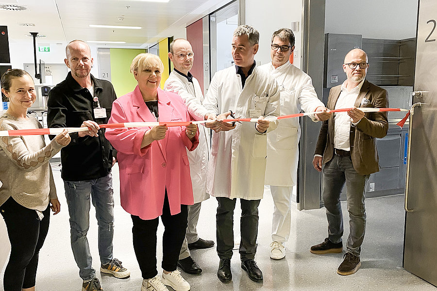 Eine Gruppe von sieben Personen steht in einem Flur und hält ein rotes und weißes Absperrband. Die Personen tragen unterschiedliche Kleidung, darunter ein weißer Kittel und eine pinke Jacke. Im Hintergrund ist eine Tür mit der Nummer '24' sichtbar.