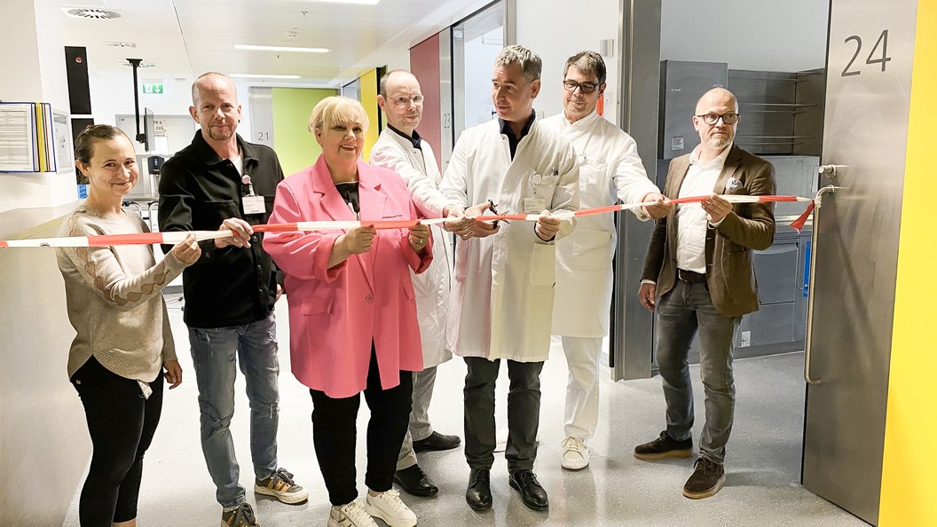 Eine Gruppe von sieben Personen steht in einem Flur und hält ein rotes und weißes Absperrband. Die Personen tragen unterschiedliche Kleidung, darunter ein weißer Kittel und eine pinke Jacke. Im Hintergrund ist eine Tür mit der Nummer '24' sichtbar.