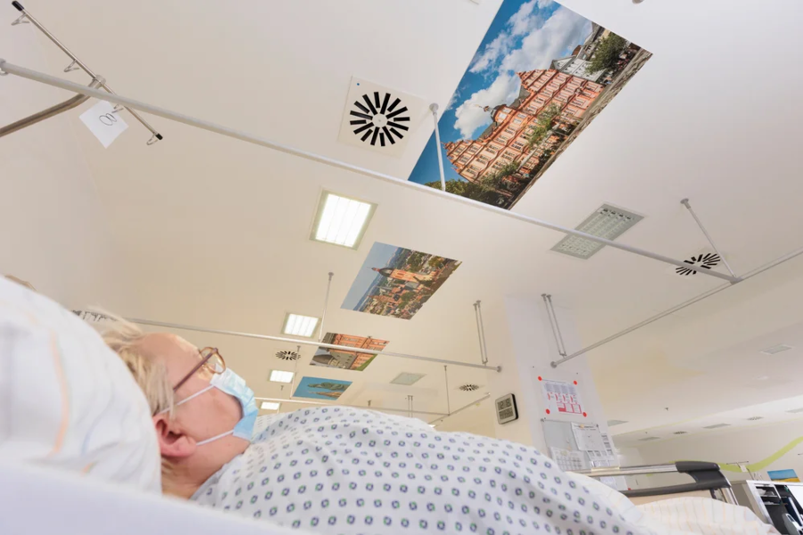 Eine Patientin liegt im Bett und schaut nach oben. An der Decke sind mehrere Bilder von Gebäuden angebracht. Die Patientin trägt eine Maske und hat kurzes, blondes Haar.