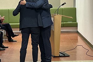 Zwei Männer in Anzügen umarmen sich vor einem Rednerpult in einem Raum mit grünen Sitzbänken