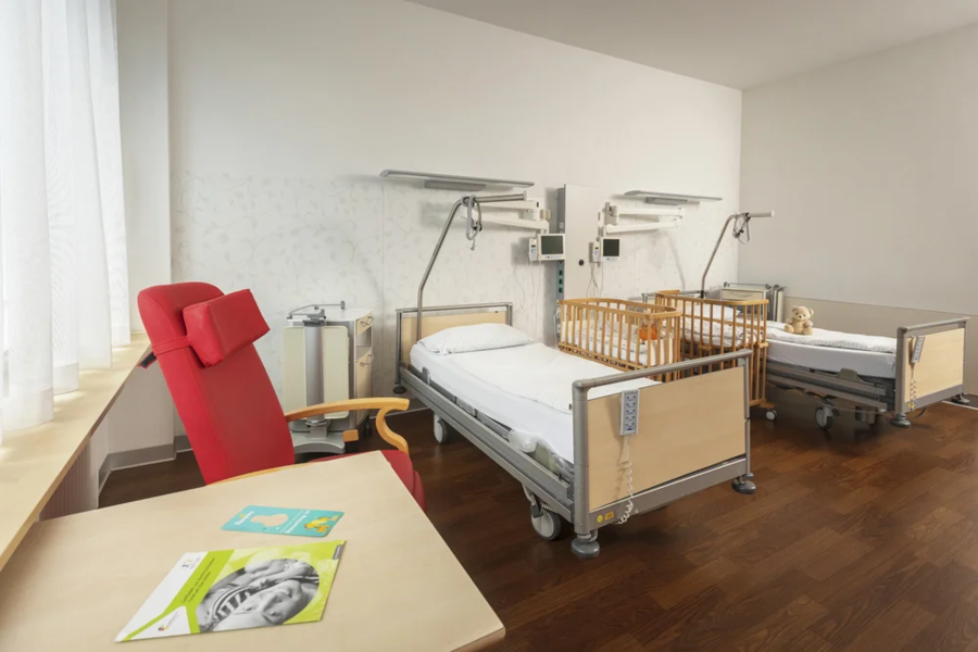 Ein Krankenhauszimmer mit zwei Pflegebetten, einem Kinderbett und einem roten Sessel. Auf dem Tisch liegen ein grünes Heft und ein Bild.