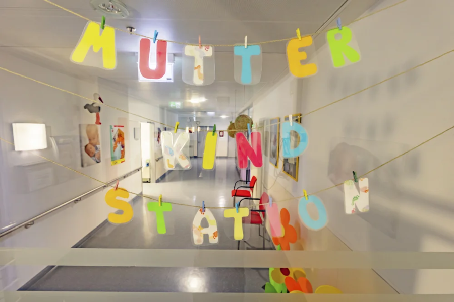 Bunte Buchstaben hängen an einer Schnur und bilden die Worte 'MUTTER KIND STATION'. Im Hintergrund sind Stühle und Bilder an der Wand sichtbar.