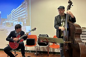 Zwei Musiker spielen in einem Raum, einer sitzt mit einer E-Gitarre, der andere steht mit einem Kontrabass, im Hintergrund eine Leinwand mit einem Bild und Text