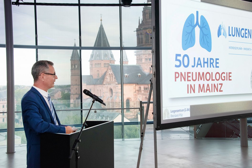 Mann im blauen Anzug steht am Rednerpult vor Mikrofon, im Hintergrund Fenster mit Blick auf Mainzer Dom. Rechts Bildschirm zeigt Text '50 Jahre Pneumologie in Mainz' und Lungen-Logo.