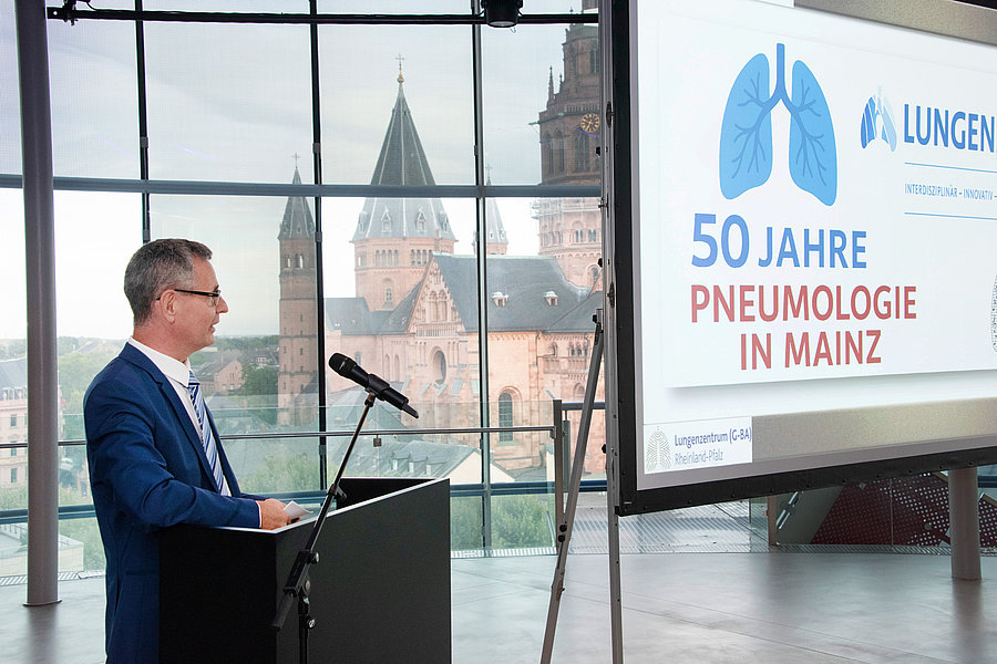 Mann im blauen Anzug steht am Rednerpult vor Mikrofon, im Hintergrund Fenster mit Blick auf Mainzer Dom. Rechts Bildschirm zeigt Text '50 Jahre Pneumologie in Mainz' und Lungen-Logo.