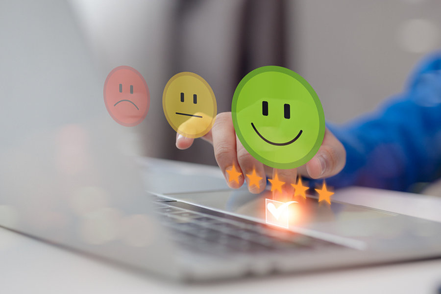 Eine Hand mit blauer Kleidung tippt auf eine Laptop-Tastatur. Über der Tastatur schweben drei emoticonartige Symbole: ein rotes trauriges Gesicht, ein gelbes neutrales Gesicht und ein grünes lächelndes Gesicht, sowie fünf goldene Sterne.