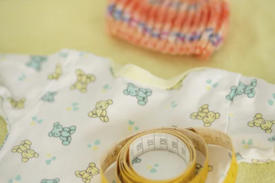Ein kleines Babyshirt mit Bärenmuster, eine orange gestrickte Mütze und ein Maßband liegen auf einer gelben Unterlage.