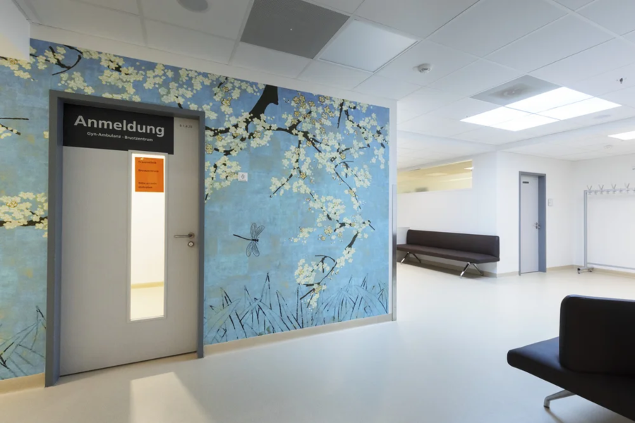 Tür mit dem Schild 'Anmeldung' in einem Raum mit blühenden Kirschblüten an der Wand. Im Hintergrund eine Bank und ein Kleiderständer.