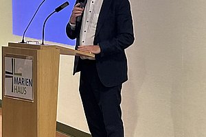 Mann in Anzug steht an Rednerpult mit Mikrofon und hält weiteres Mikrofon in der Hand