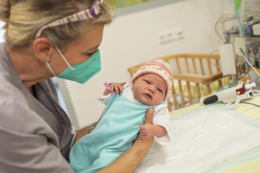 Eine Krankenschwester hält ein Baby in einem blauen Strampler mit weißen Ärmeln. Im Hintergrund ist ein Krankenhausbett und medizinische Geräte sichtbar.