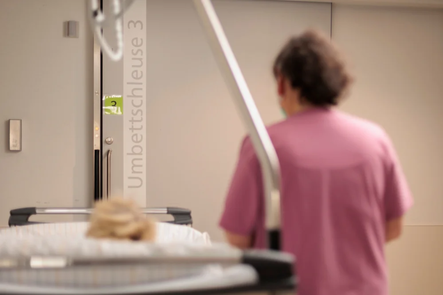 Krankenhausflur mit Patient auf Liege und medizinischem Personal in rosa Kleidung, Schild 'Umbettschleuse 3' an der Wand
