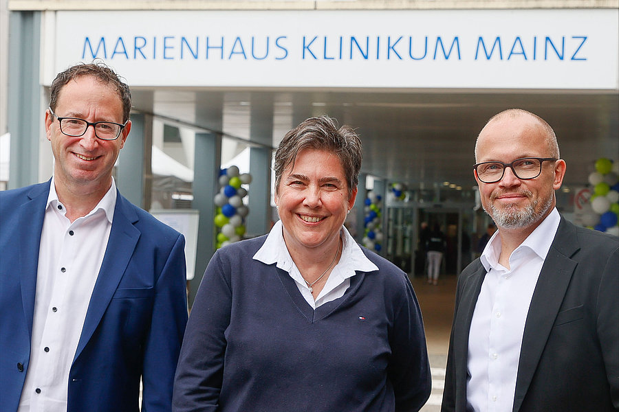 Drei Personen stehen vor dem Eingang des Marienhaus Klinikums Mainz, erkennbar am Schriftzug über dem Eingang.