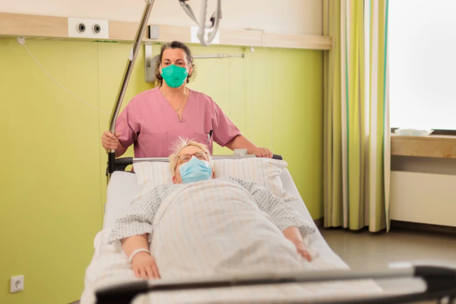 Krankenpflegerin in rosa Kittel schiebt Patientin auf Krankenhausbett durch einen hellen Raum mit grüner Wand