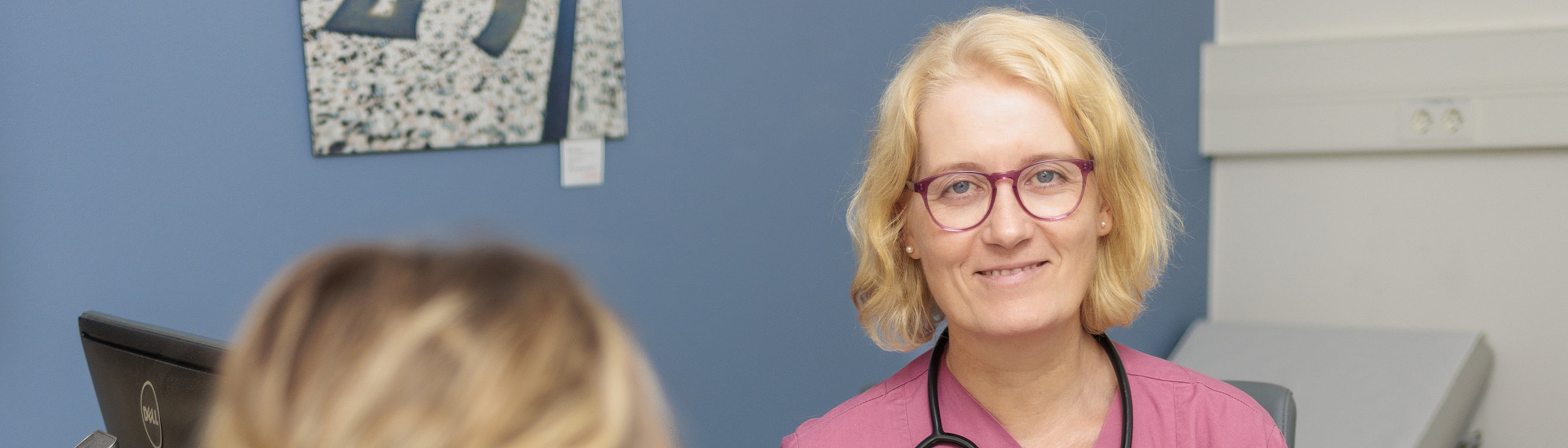 Ärztin in Rosa mit Stethoskop am Tisch, Gespräch mit Patientin in schwarzer Kleidung.