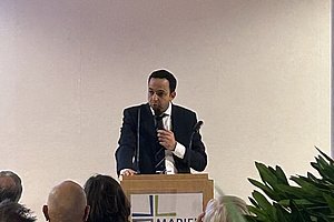 Mann in Anzug steht hinter einem Rednerpult und spricht vor Publikum in einem hellen Raum