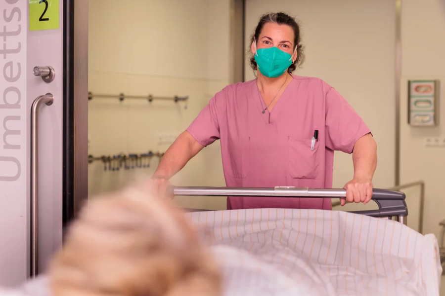 Pflegekraft in rosa Kittel schiebt Krankenhausbett durch Flur