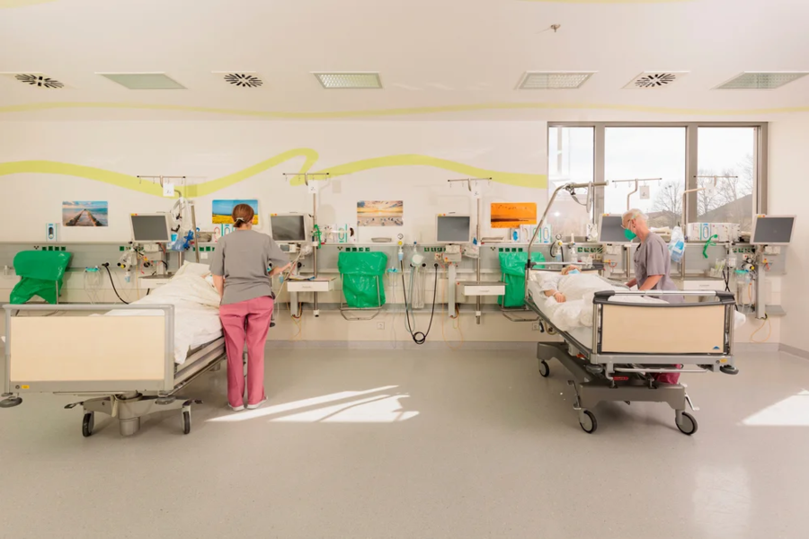 Krankenhauszimmer mit zwei Patientenbetten, medizinischen Geräten und zwei Pflegekräften in Schutzkleidung