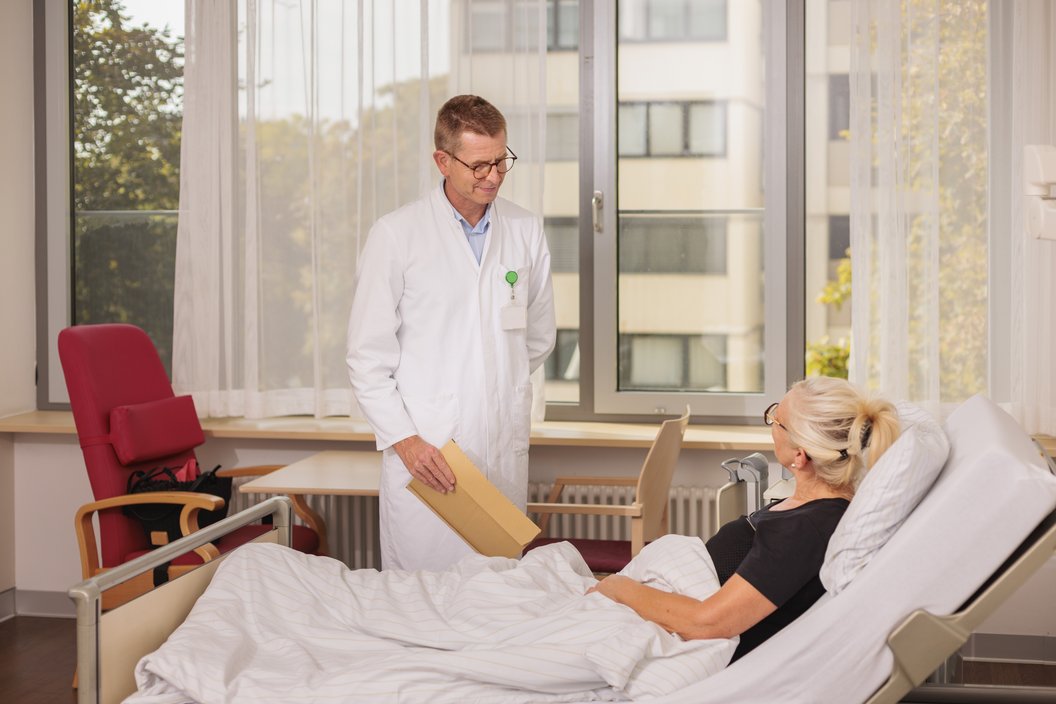 Ein Arzt in einem weißen Kittel steht neben einem Bett mit einer Patientin. Der Arzt hält eine braune Mappe in der Hand. Im Hintergrund sind Fenster mit Tageslicht und ein roter Stuhl sichtbar.
