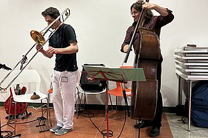 Zwei Musiker spielen in einem Raum, einer mit Posaune, der andere mit Kontrabass, vor einem weißen Hintergrund mit gestapelten Stühlen