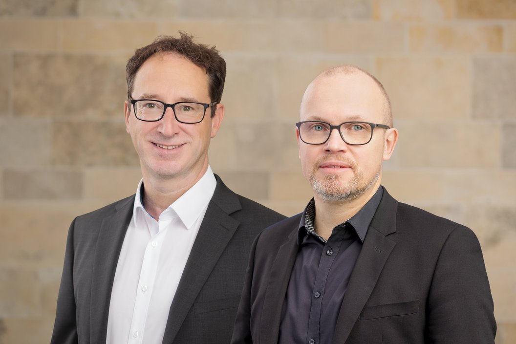 Herr Link und Herr Nietzel stehen nebeneinander, beide tragen schwarze Anzüge, Herr Link ein weißes Hemd, Herr Nietzel ein schwarzes Hemd