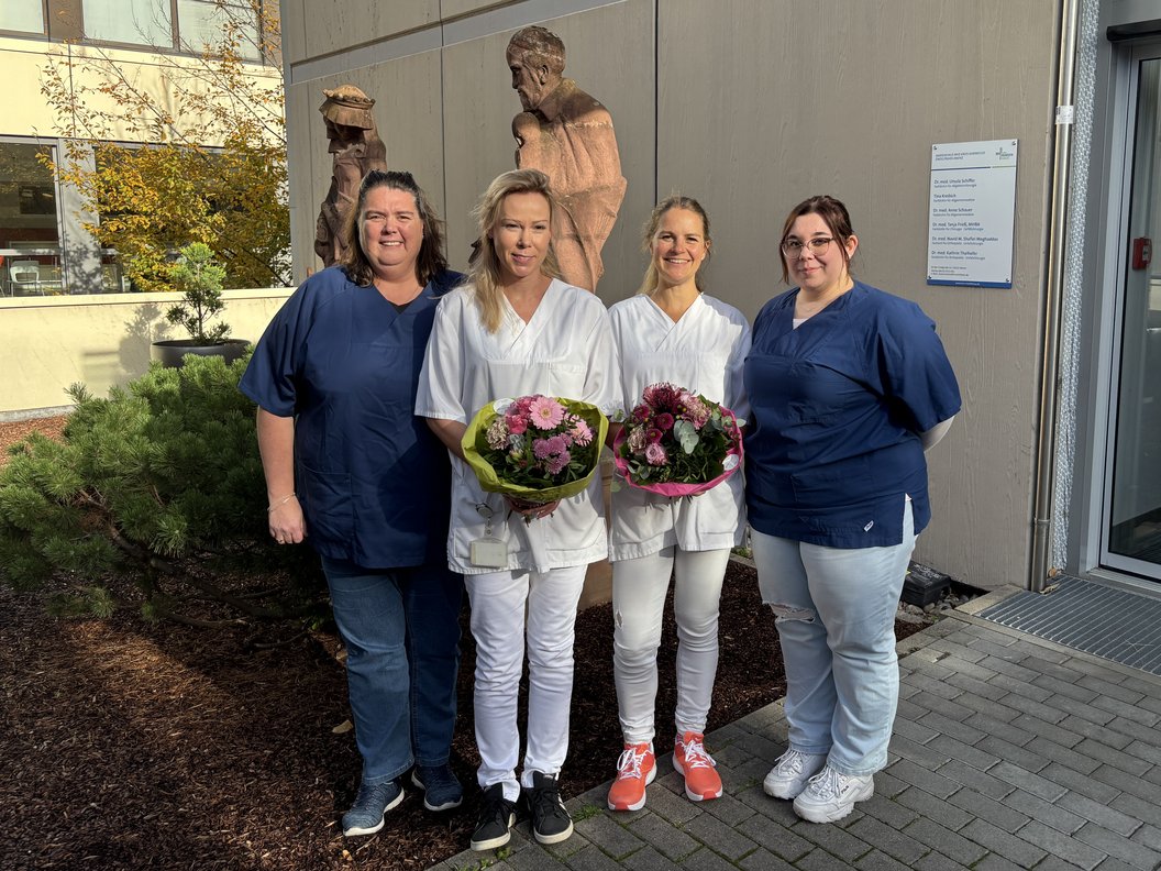 Fünf Personen in medizinischer Arbeitskleidung stehen vor einem Gebäude, zwei in weißer und drei in blauer Kleidung, zwei halten Blumensträuße.