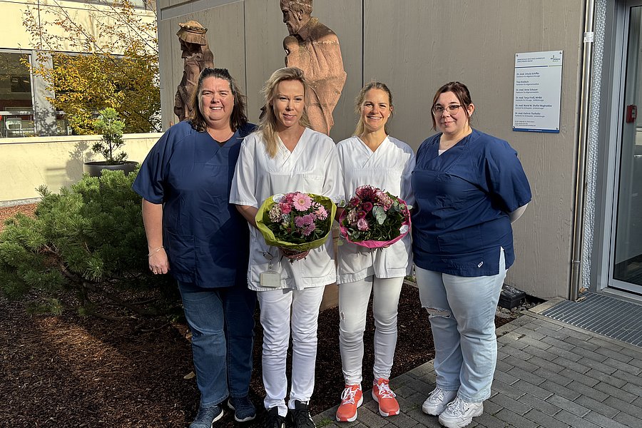 Fünf Personen in medizinischer Arbeitskleidung stehen vor einem Gebäude, zwei in weißer und drei in blauer Kleidung, zwei halten Blumensträuße.