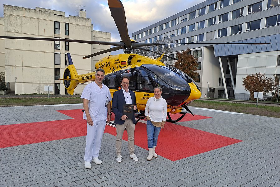 Drei Personen stehen vor einem gelb-schwarzen ADAC-Luftrettungshubschrauber auf einem gepflasterten Landeplatz mit rotem Markierungsfeld.