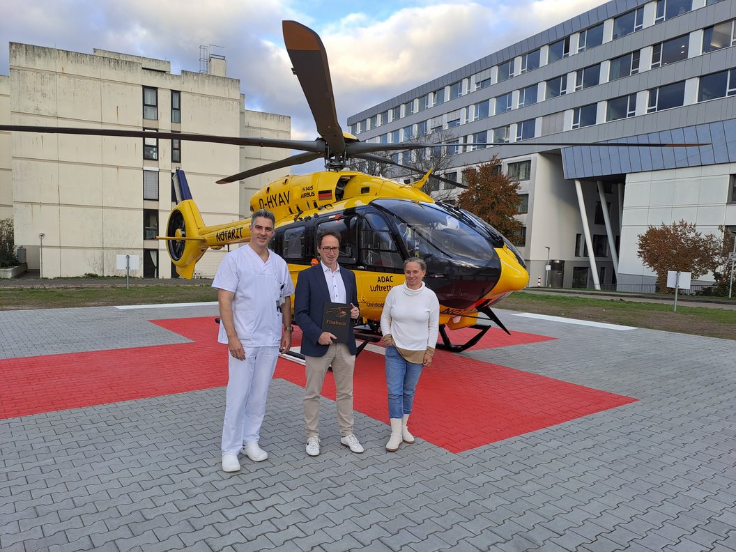 Drei Personen stehen vor einem gelb-schwarzen ADAC-Luftrettungshubschrauber auf einem gepflasterten Landeplatz mit rotem Markierungsfeld.