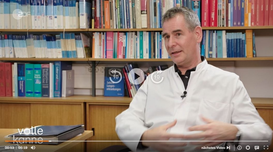 Ein Mann in einem weißen Kittel sitzt in einem Büro und gestikuliert mit beiden Händen. Im Hintergrund sind Regale mit bunten Büchern und Ordnern zu sehen.